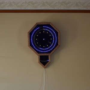 May include: A wooden octagonal wall clock with a blue and white LED light display. The clock is hanging on a beige wall with a white cord plugged into an outlet. There is a brown leather armchair with a black and white throw blanket in the foreground.