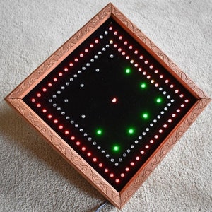 May include: A square LED light display with a copper-coloured frame and a black interior. The display features red, green, and white lights arranged in a pattern. The frame has a decorative carved design. The light is plugged in with a grey cord.