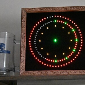 May include: A wooden framed clock with a black background and a circular design of red, green, and silver lights. The clock is on a shelf with a glass mug that says "Penn State" on it.