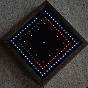 May include: A square, framed artwork with a black interior, lit by many small LED lights. The lights are arranged in a diamond pattern, with white, red, and yellow lights. The frame is a dark brown colour with an ornate design.
