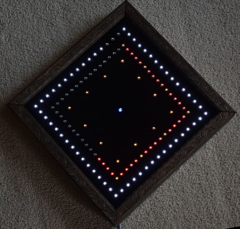 May include: A square, framed artwork with a black interior, lit by many small LED lights. The lights are arranged in a diamond pattern, with white, red, and yellow lights. The frame is a dark brown colour with an ornate design.