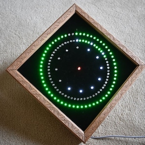 Puede incluir: Un reloj cuadrado con fondo negro y una pantalla LED verde y blanca. El reloj está enmarcado en madera con un diseño de hojas talladas.