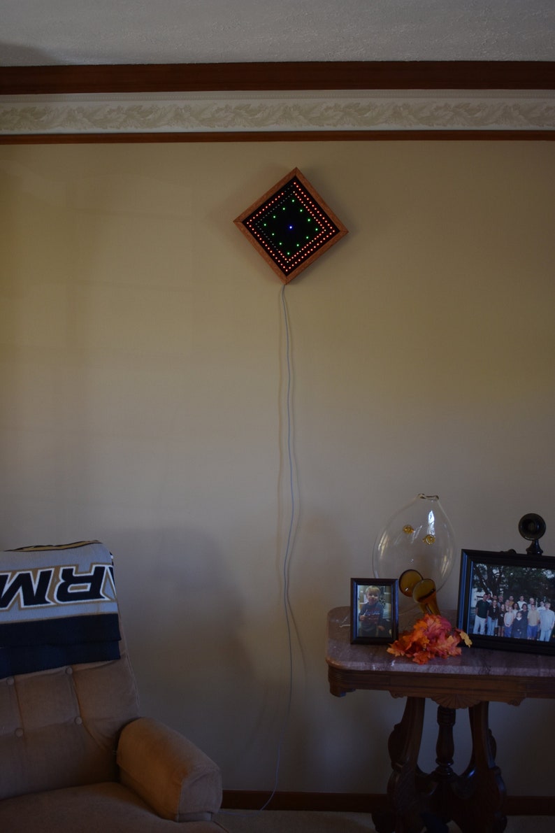 May include: A diamond-shaped LED light fixture with a wooden frame, featuring red, green, and blue lights, is mounted on a beige wall. A power cord hangs down. A side table holds framed photos and a decorative glass vase.