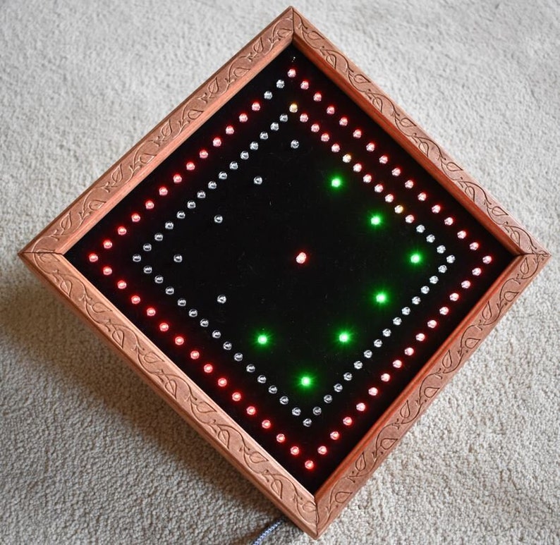 May include: A square LED light display with a copper-coloured frame and a black interior. The display features red, green, and white lights arranged in a pattern. The frame has a decorative carved design. The light is plugged in with a grey cord.