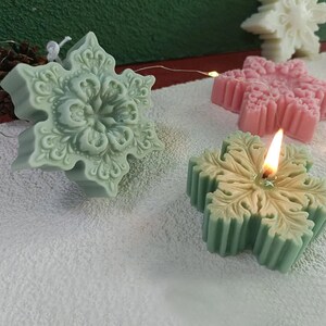 May include: Three snowflake-shaped candles, one lit, in green, pink, and white. The candles are on a white surface.