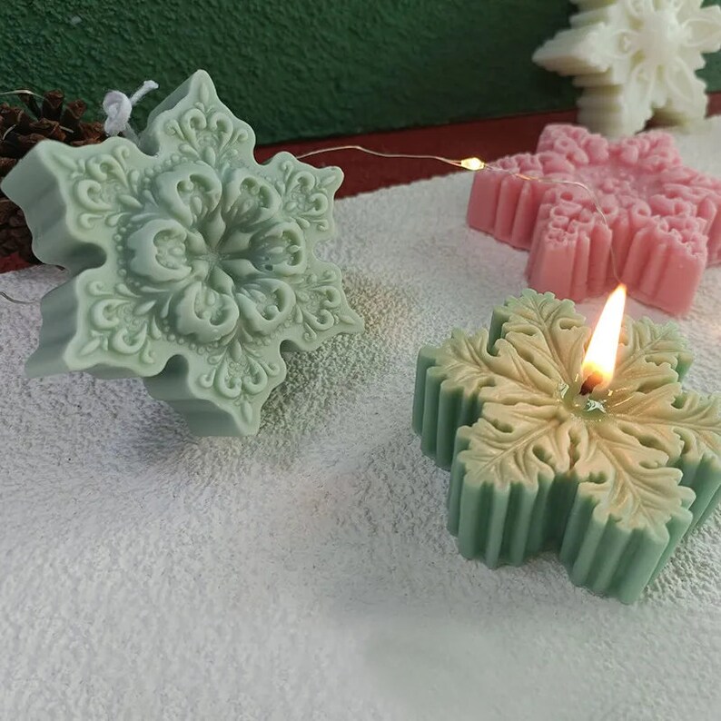 May include: Three snowflake-shaped candles, one lit, in green, pink, and white. The candles are on a white surface.