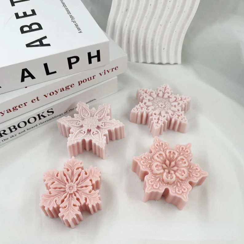 May include: Four pink snowflake-shaped soaps with intricate designs. The soaps are arranged on a white surface.