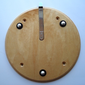 Puede incluir: Un soporte de pared redondo de madera con un gancho de metal y cuatro pies de goma negra. El soporte está hecho de madera clara con un acabado natural.