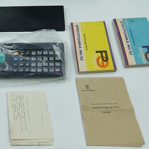 May include: A black calculator with a green display and a black case. The calculator is in a plastic bag. There are two instruction manuals with the text "Электроника МК-52" and a brown passport-sized booklet with the text "Электроника ВЛ-37А" on it.