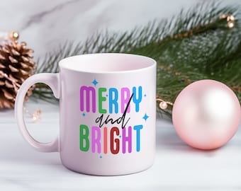 Könnte beinhalten: Eine hellrosa Keramik-Tasse mit den Worten "MERRY and BRIGHT" in bunten Blockbuchstaben. Die Tasse hat einen Henkel und ist mit blauen Sternen verziert. Eine rosa Kugel und ein Tannenzapfen sind im Hintergrund.