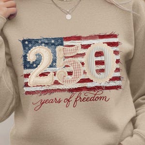 May include: Beige sweatshirt with a distressed American flag design. The flag features the number "250" in a patchwork style, with the words "years of freedom" in script below. The sweatshirt is a neutral color.