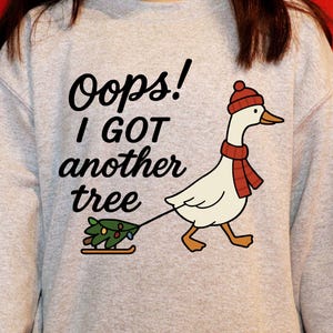May include: A light gray sweatshirt with the text "Oops! I GOT another tree." A cartoon goose wearing a red hat and scarf pulls a small Christmas tree on a sled. The sweatshirt is a long-sleeved crew neck.