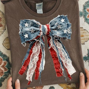 May include: A dark brown t-shirt featuring a large, decorative bow graphic. The bow is constructed from distressed blue denim fabric adorned with white stars, layered with frayed red fabric strips and delicate white lace trim accents.