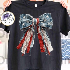 May include: Black t-shirt featuring a distressed denim bow design with red, white, and blue accents. The bow has star embellishments and lace detailing, creating a patriotic and vintage aesthetic. The shirt is held up by a person.