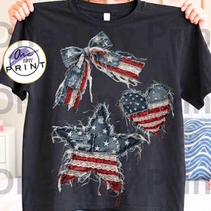 May include: A black t-shirt with patriotic designs. The shirt features a bow, a heart, and a star, each made with a distressed American flag pattern. The designs use red, white, and blue colours, creating a vintage look. The shirt is held up by a person.