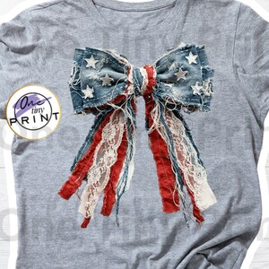 May include: A heather gray t-shirt featuring a large graphic of a distressed denim bow with white stars, red fabric strips, and white lace trim. The graphic includes a circular logo with the text One tiny PRINT on the left side of the shirt.
