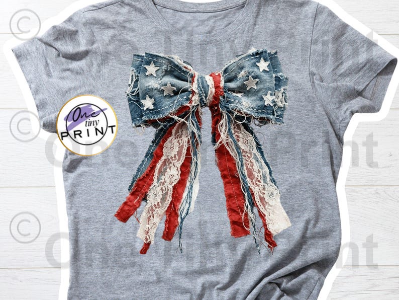 May include: A heather gray t-shirt featuring a large graphic of a distressed denim bow with white stars, red fabric strips, and white lace trim. The graphic includes a circular logo with the text One tiny PRINT on the left side of the shirt.