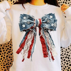 May include: A white crewneck sweatshirt featuring a large, central graphic of a bow made from distressed blue denim fabric with white star patterns, accented by frayed red fabric strips and white lace trim hanging down from the bow center.