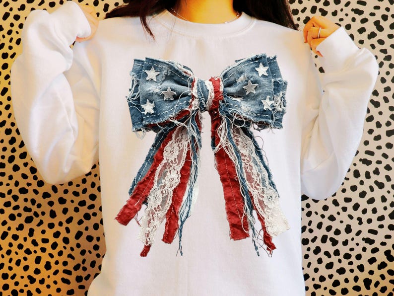 May include: A white crewneck sweatshirt featuring a large, central graphic of a bow made from distressed blue denim fabric with white star patterns, accented by frayed red fabric strips and white lace trim hanging down from the bow center.