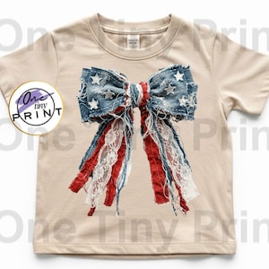 May include: A beige short-sleeve t-shirt featuring a large, centered graphic of a distressed denim bow with white stars, red fabric strips, and white lace ribbons. A circular logo on the left side contains the text One tiny PRINT.