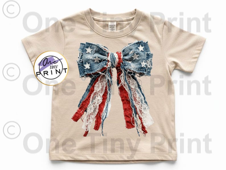 May include: A beige short-sleeve t-shirt featuring a large, centered graphic of a distressed denim bow with white stars, red fabric strips, and white lace ribbons. A circular logo on the left side contains the text One tiny PRINT.