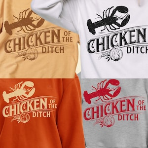 Puede incluir: Cuatro camisetas y sudaderas en varios colores, cada una con la frase "Chicken of the Ditch" con un gráfico de langosta. Los colores incluyen beige, blanco, naranja y gris.
