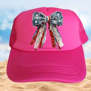 May include: A bright pink trucker hat featuring a central graphic of a distressed denim bow. The bow is decorated with small white stars and includes hanging strips of red fabric, white lace, and frayed blue denim material on the front panel.