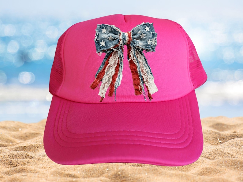 May include: A bright pink trucker hat featuring a central graphic of a distressed denim bow. The bow is decorated with small white stars and includes hanging strips of red fabric, white lace, and frayed blue denim material on the front panel.
