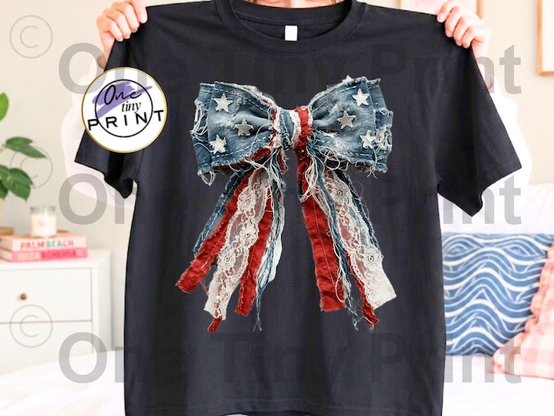 May include: Black t-shirt featuring a distressed denim bow design with red, white, and blue accents. The bow has star embellishments and lace detailing, creating a patriotic and vintage aesthetic. The shirt is held up by a person.