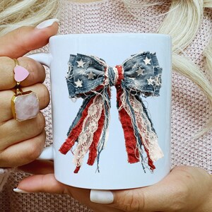 May include: A white ceramic mug featuring a graphic print of a decorative bow. The bow is constructed from frayed denim fabric with white star patterns, layered with strips of red fabric and cream-colored lace, creating a textured appearance.