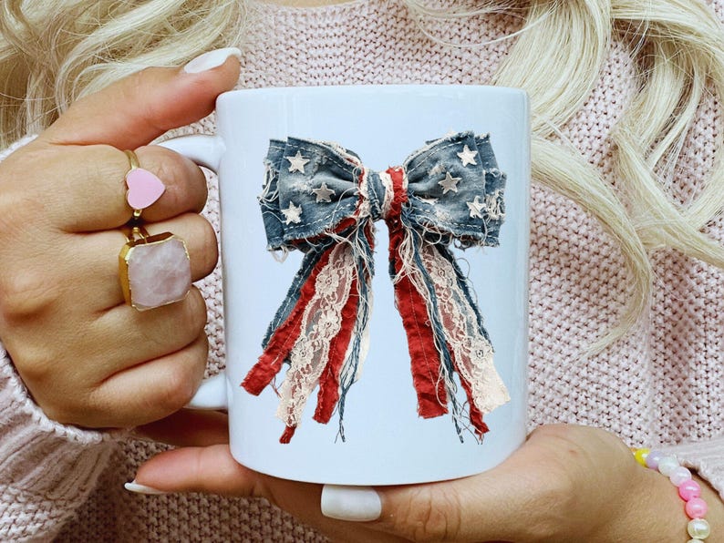 May include: A white ceramic mug featuring a graphic print of a decorative bow. The bow is constructed from frayed denim fabric with white star patterns, layered with strips of red fabric and cream-colored lace, creating a textured appearance.