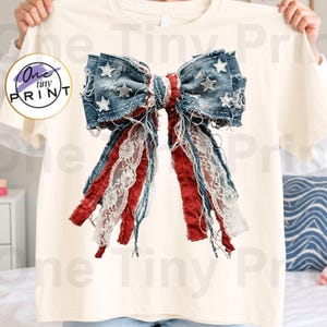 May include: A cream-colored t-shirt featuring a large, central graphic of a distressed denim bow. The bow is adorned with white stars and incorporates frayed strips of red fabric and white lace, creating a textured, layered appearance.