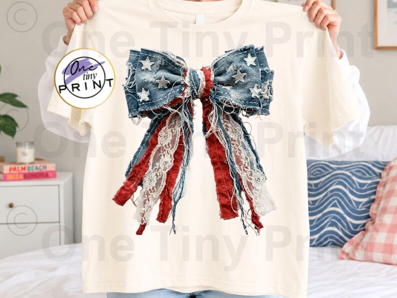 May include: A cream-colored t-shirt featuring a large, central graphic of a distressed denim bow. The bow is adorned with white stars and incorporates frayed strips of red fabric and white lace, creating a textured, layered appearance.