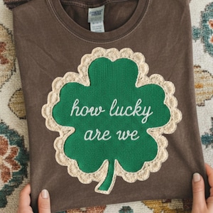 以下が含まれることがあります： 緑のクローバーのデザインと白い文字「how lucky are we」が特徴の茶色のTシャツ。クローバーはクリーム色のスカラップトリムで縁取られています。シャツは柔らかい素材で作られています。