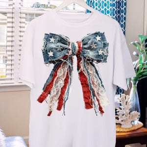May include: A white short-sleeve t-shirt featuring a large, centered graphic of a decorative bow. The bow is constructed from distressed blue denim fabric with white star patterns, accented by frayed red fabric strips and white lace ribbons.