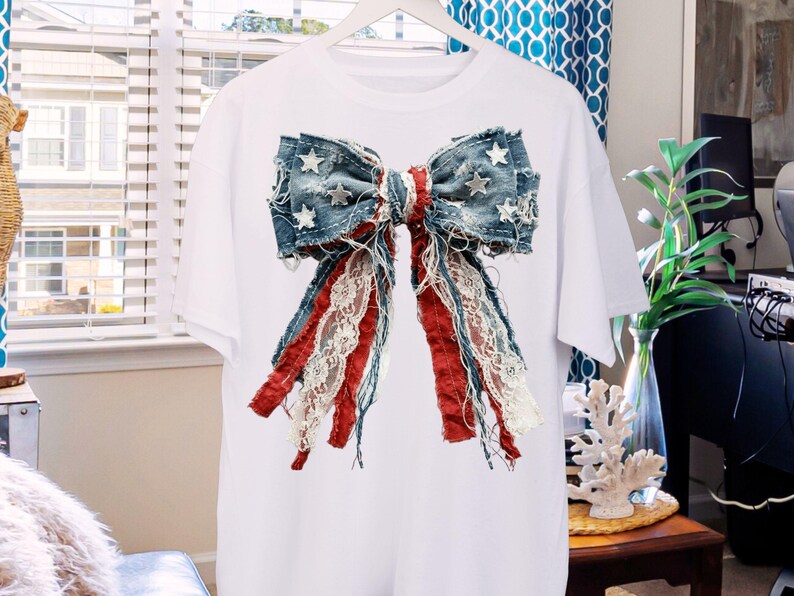 May include: A white short-sleeve t-shirt featuring a large, centered graphic of a decorative bow. The bow is constructed from distressed blue denim fabric with white star patterns, accented by frayed red fabric strips and white lace ribbons.