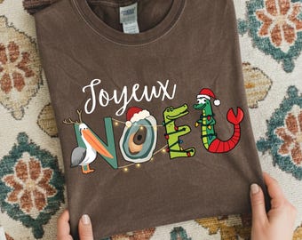 Op de afbeelding: Bruin t-shirt met de Franse uitdrukking "Joyeux Noel" in witte letters. Het woord "NOEL" is creatief ontworpen met een pelikaan, een oester en twee alligator-zeemeermin hybriden, allemaal met kerstmutsen en feestverlichting.