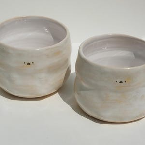 Op de afbeelding: Twee handgemaakte keramische kommen met een afgeronde vorm en een crèmewitte glazuur. Elke kom heeft een klein, minimalistisch gezichtsontwerp. De kommen hebben een licht gestructureerd oppervlak en een lichtbeige tint op sommige plaatsen.