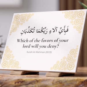 May include: A white decorative tile with gold floral corner designs and Arabic calligraphy. The English text reads, "Which of the favors of your lord will you deny?" from Surah Ar-Rahman (55:13). The tile is displayed on a wooden stand.
