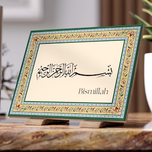 May include: A decorative framed art piece with Arabic calligraphy and the English word "Bismillah". The frame has a green border with floral patterns, and the artwork is displayed on a wooden stand. The background is neutral, with candles and a plant.