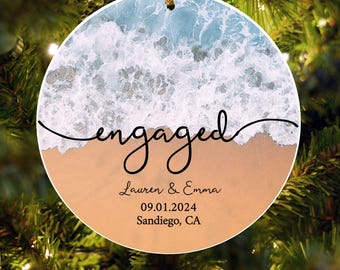 May include: A round, white, and blue Christmas ornament with a beach scene and the word "engaged" in black cursive writing. The ornament also includes the text "Lauren & Emma 09.01.2024 Sandiego, CA".