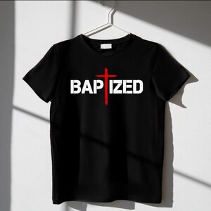May include: Black t-shirt with the word "BAPTIZED" in white block letters and a red cross graphic. The shirt is hanging on a white hanger against a white wall, with shadows cast across the surface.