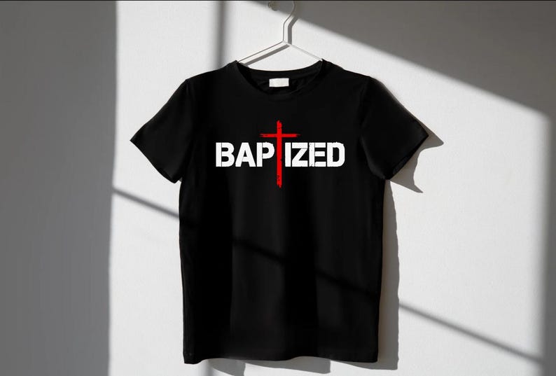 May include: Black t-shirt with the word "BAPTIZED" in white block letters and a red cross graphic. The shirt is hanging on a white hanger against a white wall, with shadows cast across the surface.