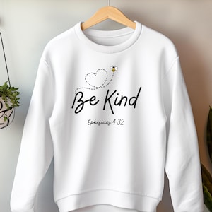 May include: White sweatshirt with the words "Be Kind" in a handwritten font, accompanied by a dotted heart and a small bee graphic. The text "Ephesians 4:32" is below the main text. The sweatshirt is hanging on a wooden hanger.