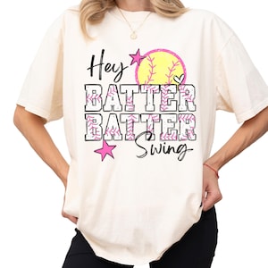 May include: Cream-colored t-shirt with the text "Hey Batter Batter Swing" in black, pink, and yellow. The design includes a softball and stars. The shirt is a casual, oversized fit.