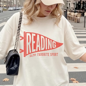 Puede incluir: Camiseta color crema con el texto "READING is my FAVORITE SPORT" en rojo y blanco, dentro de un diseño de banderín. La camiseta se combina con un bolso negro y una gorra de béisbol beige.