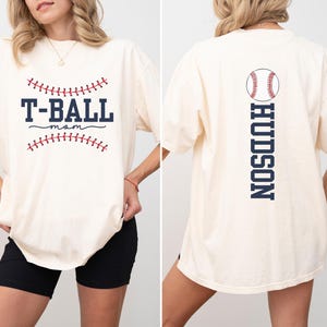 Könnte beinhalten: Ein weißes T-Shirt mit einem Baseball-Motiv und dem Text "T-BALL mom" auf der Vorderseite. Die Rückseite des Shirts hat ein Baseball-Motiv und den Text "HUDSON" vertikal auf der Rückseite.