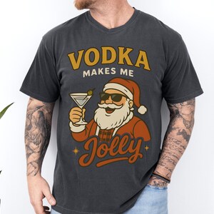 May include: Dark gray t-shirt with a graphic of Santa Claus wearing sunglasses and holding a martini. The text "VODKA makes me Jolly" is in gold and orange. A fun, festive design for the holiday season.