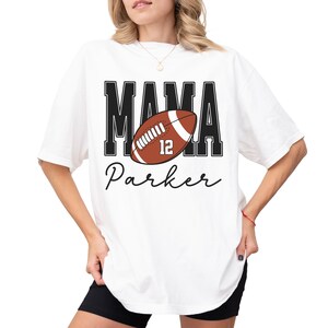 Custom Football Mama Shirt. The tshirt says football mama in varsity font and is personalized with the players number on a large football and name below it. Comfort Colors tee, crewneck, short sleeves football mom t shirt