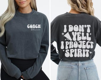May include: A dark grey long-sleeved t-shirt with the text "COACH Olivia" on the left side and "I DON'T YELL I PROJECT SPIRIT" on the right side. The text is in a white retro font with a megaphone graphic.